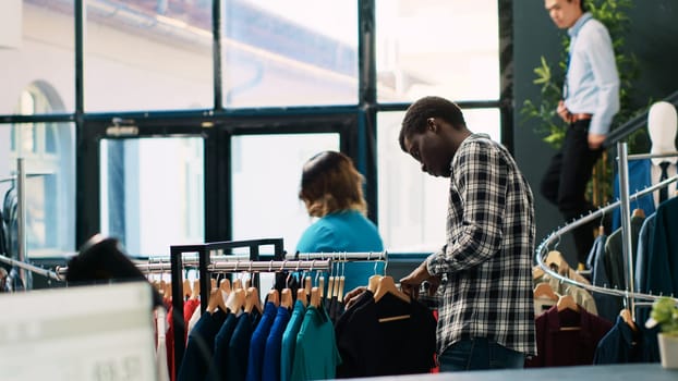 African american client shopping for black shirt, analyzing fashionable clothes material in modern boutique. Shopaholic man buying casual wear, looking at hangers with new fashion collection