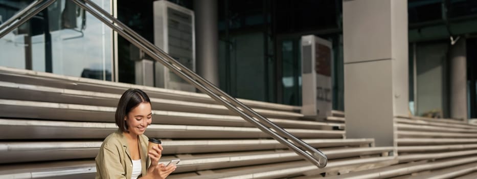Vertical shot of young smiling woman sits on stairs in city centre wth mobile phone. Girl with telephone, browsing on smartphone, texting message.
