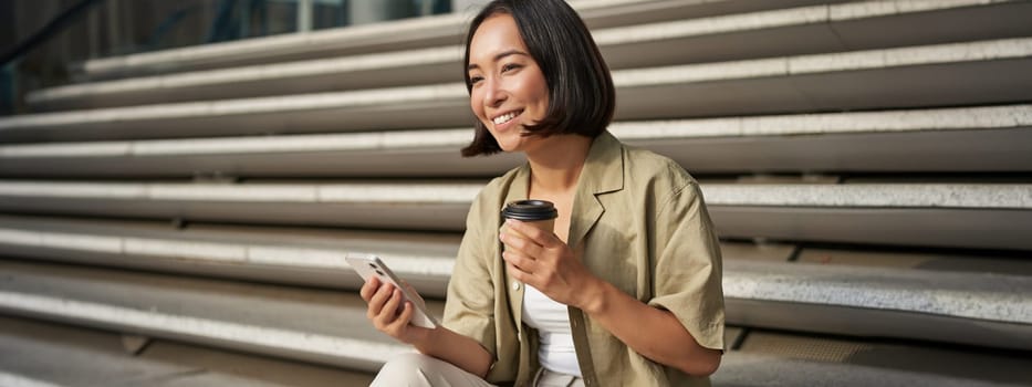 Portrait of asian woman with smartphone, drinks coffee and watches videos on mobile phone. Girl with telephone sits on stairs outdoors.