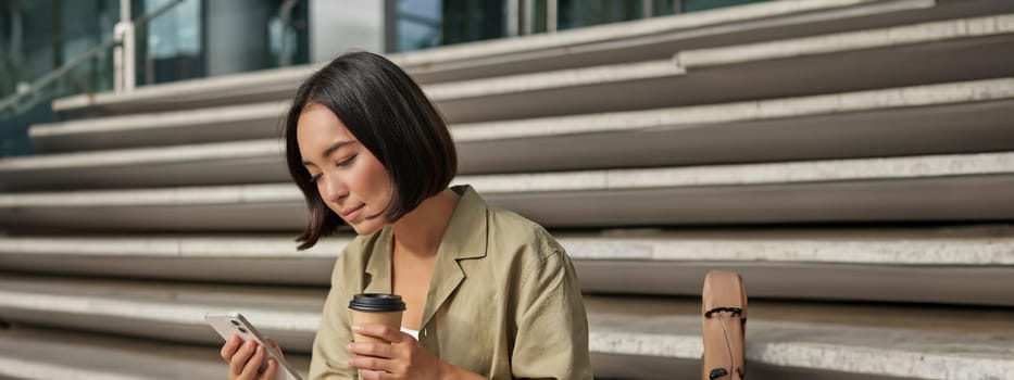 Vertical shot of asian girl drinking her coffee and using mobile phone. Young woman with smartphone and takeaway sits on stairs.