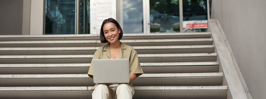 Young asian woman using laptop outdoors. Cute girl student with computer, does her homework, types, sits on stairs outside campus.