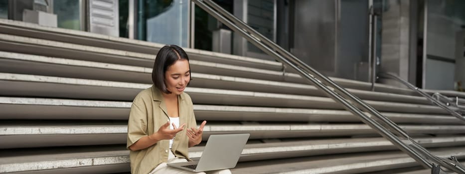 Vertical shot of asian girl student attend online meeting, talking at laptop, video chat, sitting on stairs.