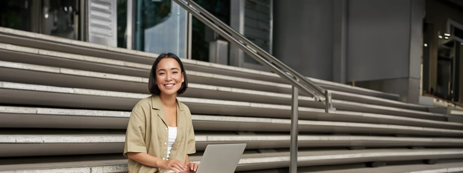 Vertical shot of asian girl sits with laptop, drinks coffee on university stairs. Young woman, student does her homework outdoors.