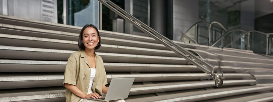 Vertical shot of asian girl sits with laptop, drinks coffee on university stairs. Young woman, student does her homework outdoors.