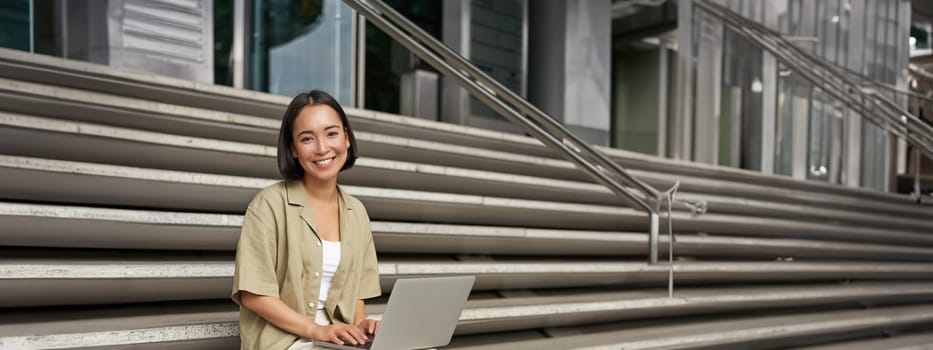 Vertical shot of asian girl sits with laptop, drinks coffee on university stairs. Young woman, student does her homework outdoors.