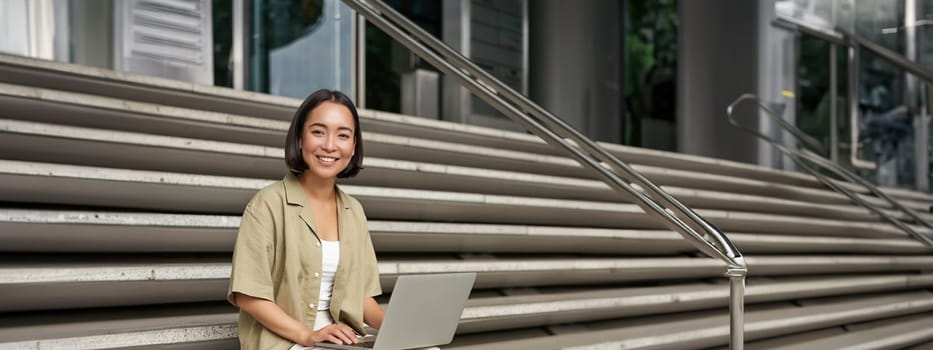 Vertical shot of asian girl sits with laptop, drinks coffee on university stairs. Young woman, student does her homework outdoors.