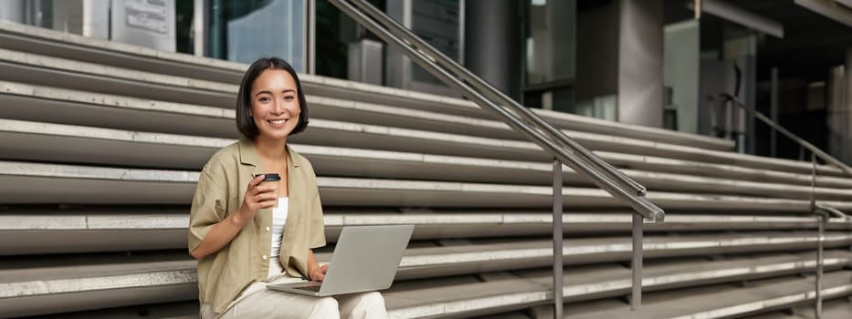 Vertical shot of smiling girl student, asian woman sits on stairs of university campus and drinks coffee, does her homework on laptop.
