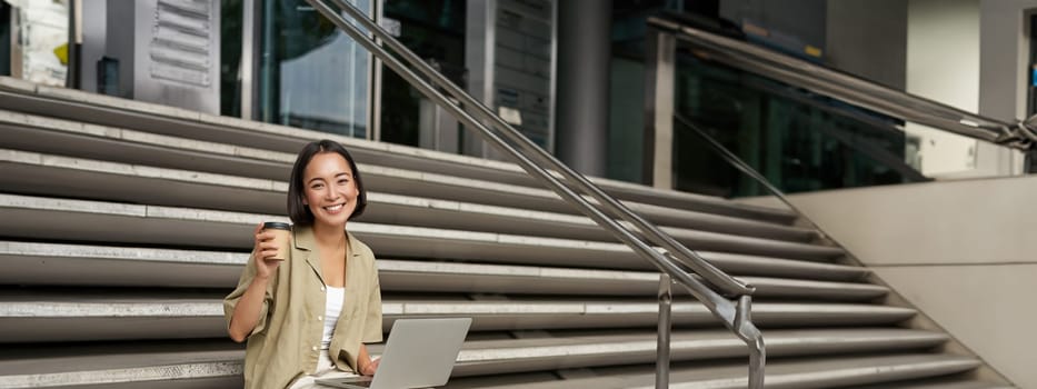 Vertical shot of smiling girl student, asian woman sits on stairs of university campus and drinks coffee, does her homework on laptop.