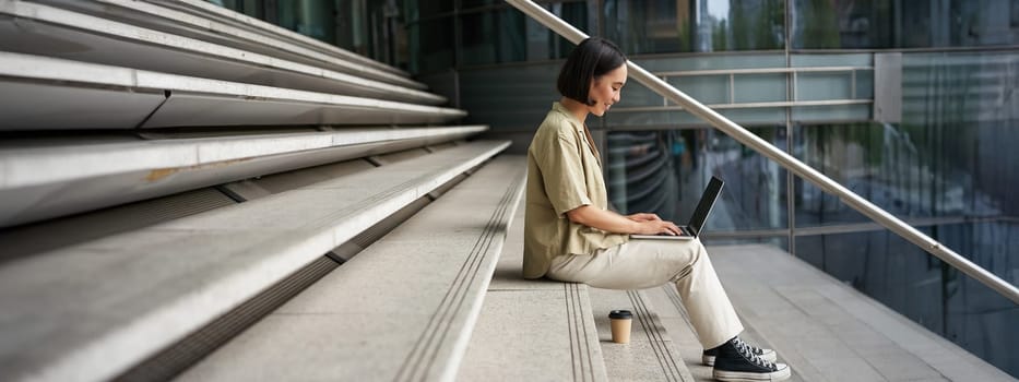 Profile portrait of young asian woman with laptop, girl student sits on stairs outside building and types on computer, drinks takeaway coffee.