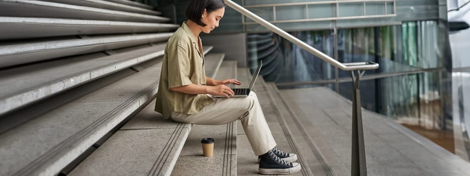 Profile portrait of young asian woman with laptop, girl student sits on stairs outside building and types on computer, drinks takeaway coffee.