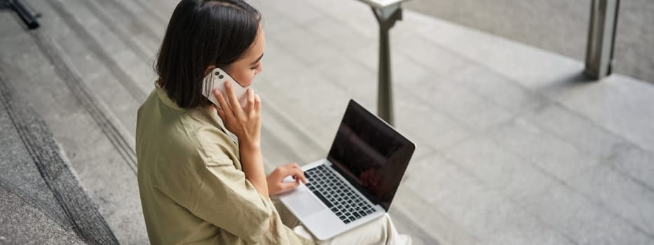 Portrait of asian girl, student sits on stairs with laptop, talks on mobile phone. Young woman makes a telephone call while working on computer.