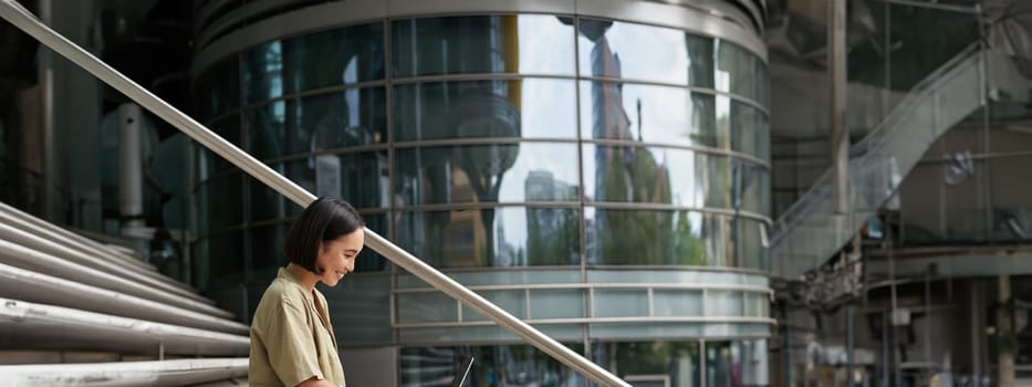 Vertical shot of smiling asian girl with coffee, uses laptop outdoors. Young woman with computer outside, sits on stairs.