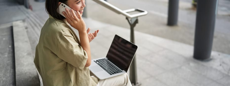 Smiling asian woman makes a phone call. Girl student using laptop and mobile phone, talking to someone on telephone.