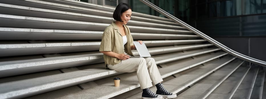 Young smiling asian girl with laptop, sits on street of city with coffee. Young woman does homework on computer while sitting on street stairs, digital nomad works remote.