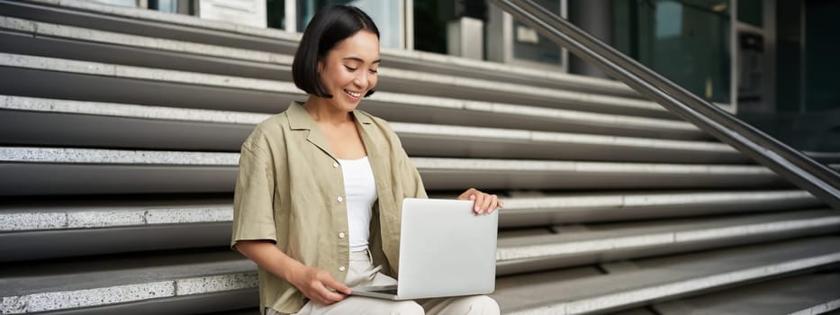 Portrait of young woman student, asian girl using laptop. Asign smiling girl, digital nomad works on her project remotely, sitting on street.