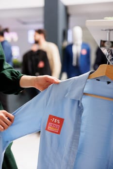Woman customer looking through clothes hanging on rack, checking Black Friday offer in trendy Fashion boutique. Male shopper holding blue shirt with 75 percent discount red sale price tag