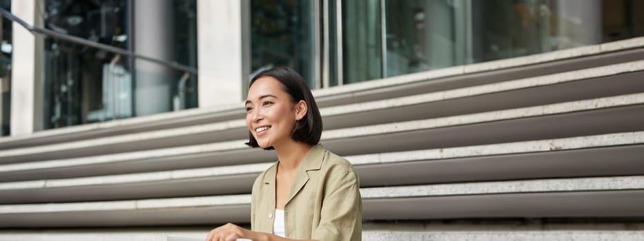 Vertical shot of smiling asian woman with laptop computer, sitting on stairs outside building. Technology and digital nomad concept
