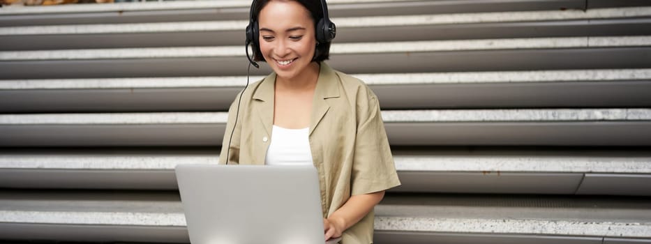 Portrait of asian woman, digital nomad girl using laptop and listens to music outdoors. Young student works on computer and smiles.
