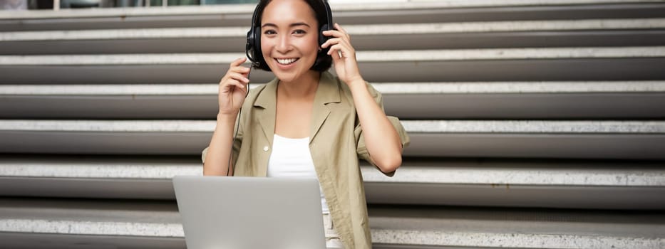 Smiling asian girl in headphones, works on laptop, digital nomad using computer on remote, sitting on stairs outdoors.