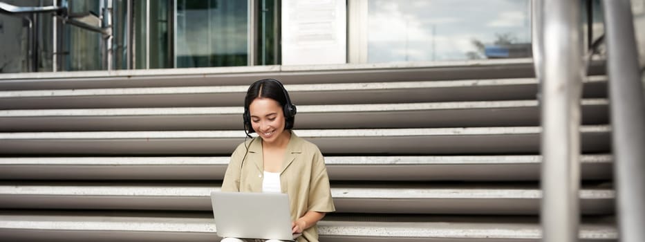 Smiling asian girl in headphones, works on laptop, digital nomad using computer on remote, sitting on stairs outdoors.