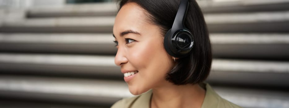 Close up portrait of smiling asian girl in headphones, listens to music outdoors, looking happy. People concept