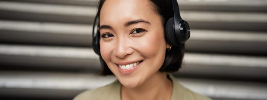 Close up portrait of smiling asian girl in headphones, listens to music outdoors, looking happy. People concept