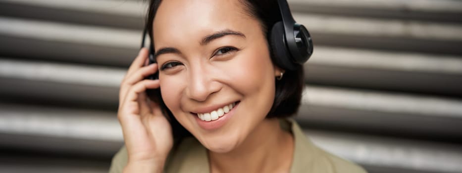 Close up portrait of smiling asian girl in headphones, listens to music outdoors, looking happy. People concept