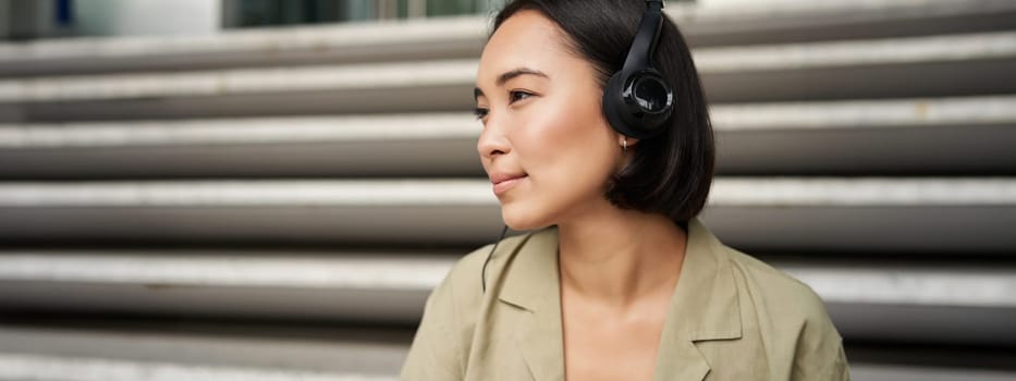 Young asian woman listening to music in headphones, sitting outside on street, smiling.