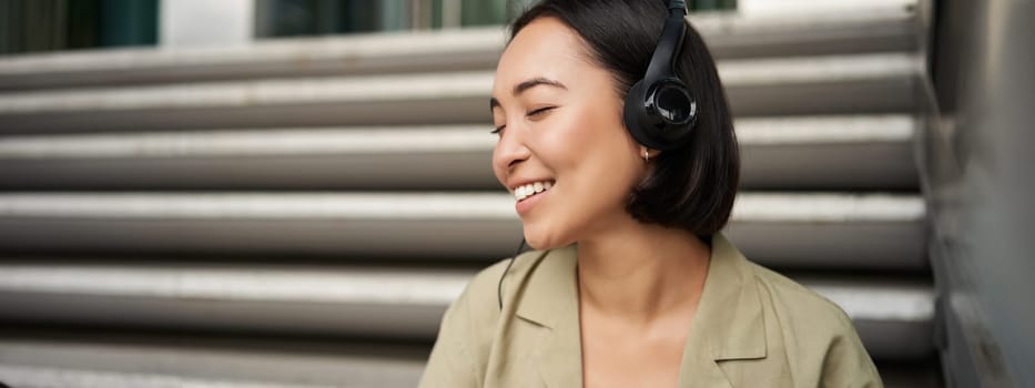 Young asian woman listening to music in headphones, sitting outside on street, smiling.