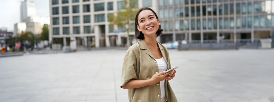 Portrait of asian woman standing on street, city square and holding mobile phone. Girl with smartphone walking outdoors.