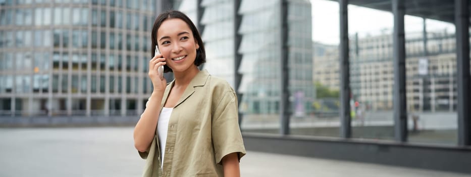 Portrait of asian girl smiling, talking on phone, making a call, standing on street near building and waiting for someone, answer telephone.