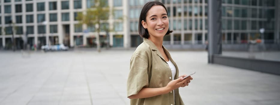 Portrait of asian woman standing on street, city square and holding mobile phone. Girl with smartphone walking outdoors.