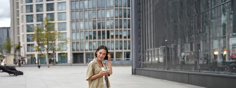 Portrait of asian woman walking in city. Street style shot of girl with smartphone, posing outdoors on street.