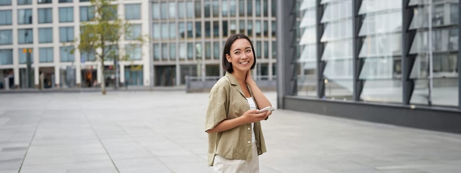 Portrait of asian woman walking in city. Street style shot of girl with smartphone, posing outdoors on street.