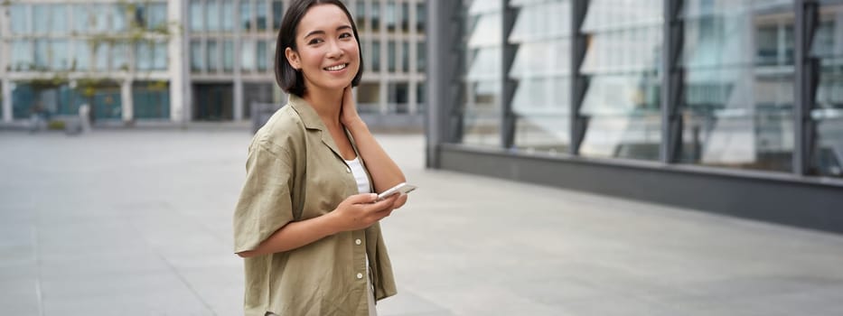 Portrait of asian woman walking in city. Street style shot of girl with smartphone, posing outdoors on street.