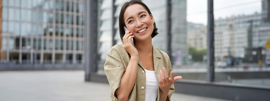 Cellular connection. Young asian woman makes a telephone call, talking on mobile smartphone and walking on street.