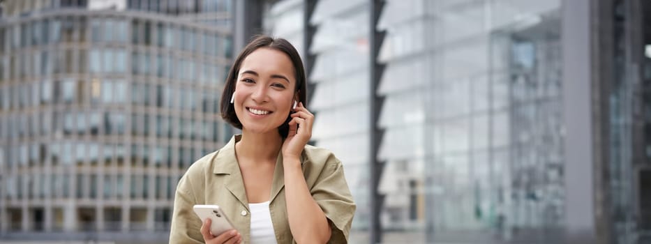 Young smiling asian woman in wireless earphones, walks on street with smartphone and headphones.