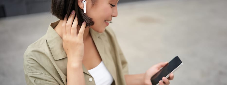 Close up portrait of asian girl, looking at mobile screen, listening music in headphones. Woman with earphones walks on street.