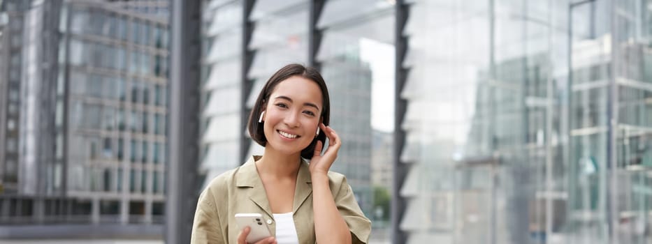 Young smiling asian woman in wireless earphones, walks on street with smartphone and headphones.