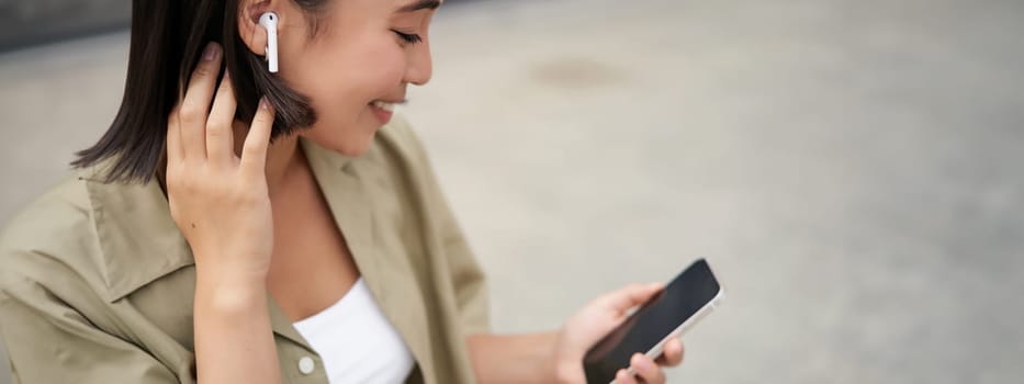 Close up portrait of asian girl, looking at mobile screen, listening music in headphones. Woman with earphones walks on street.