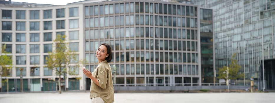 Modern people. Young woman walks on street of city and listens music in wireless headphones, holds smartphone.