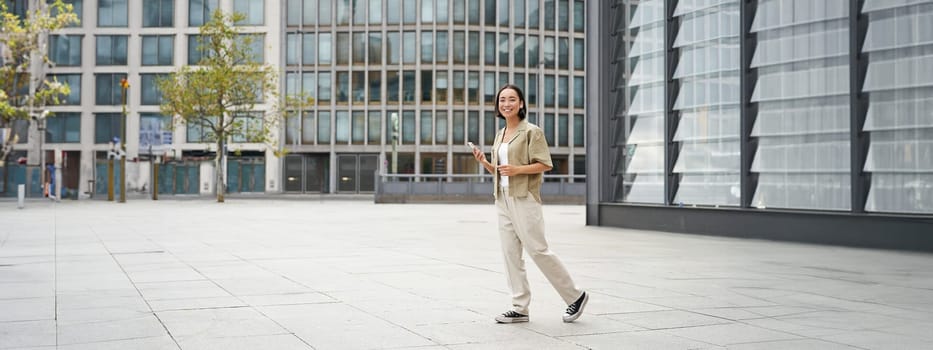 Portrait of beautiful smiling asian woman, walking on street of city centre with smartphone, looking at camera.