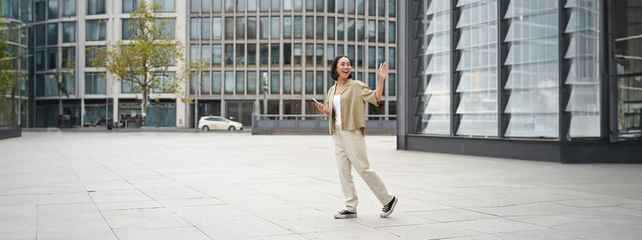 Happy asian girl saying bye to someone, waving at friend and walking on street with smartphone.