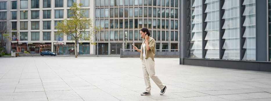 Silhouette of asian woman walking on street in wireless headphones, holding smartphone.