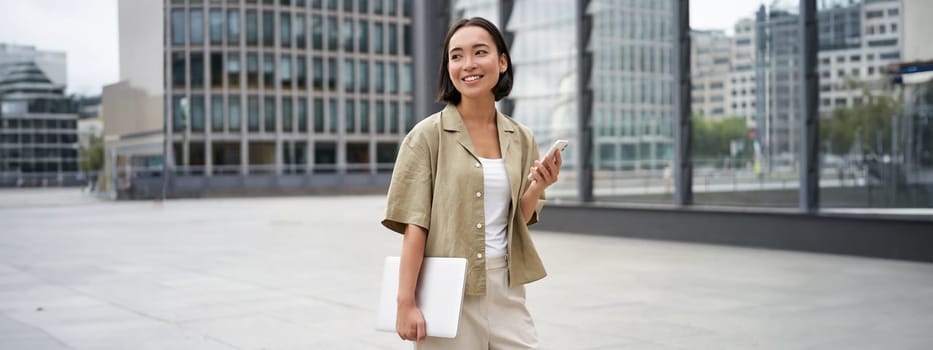 Asian girl with laptop and smartphone, standing on street of city centre, smiling at camera.