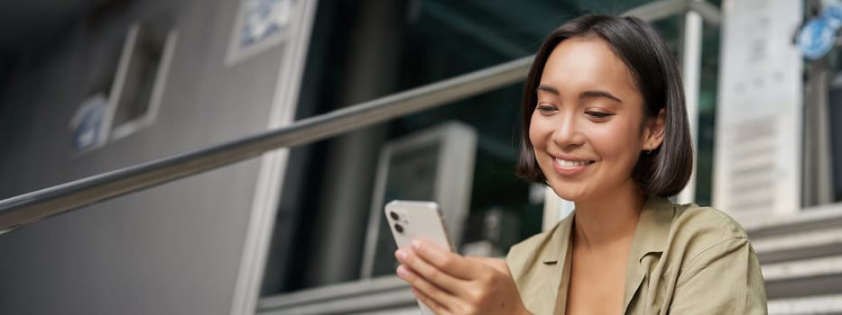 Portrait of asian girl takes selfie on mobile phone. Korean woman smiling, video chat on smartphone app while sits outside on stairs.