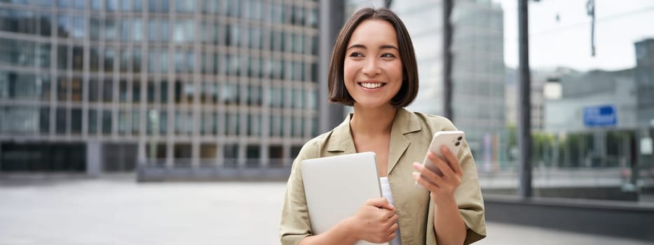 Portrait of young smiling asian woman walking on street, going to work with laptop and smartphone.