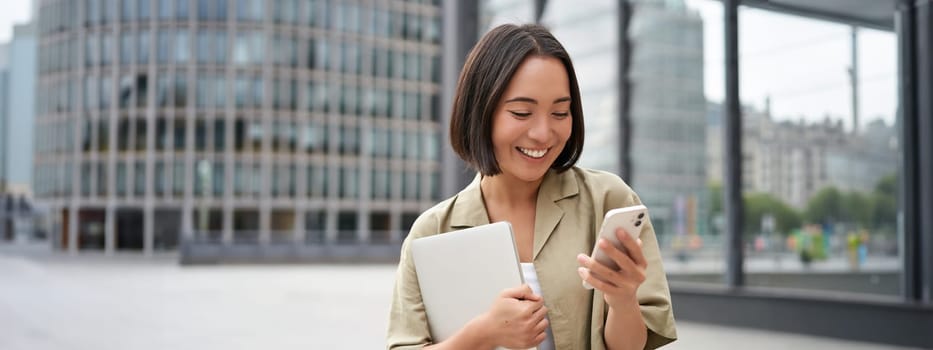 Portrait of young smiling asian woman walking on street, going to work with laptop and smartphone.