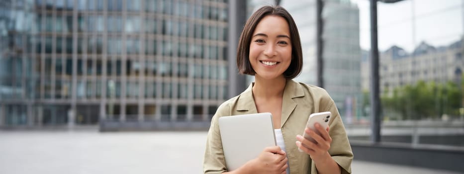 Portrait of smiling asian girl with laptop, holds mobile phone and looks happy at camera, stands on street.