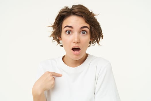 Portrait of girl with surprised face, points at herself amazed, stands isolated over white studio background. Copy space